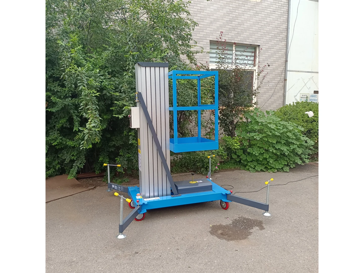 Complete blue single mast lift lowered with its stabilizers deployed outside a building