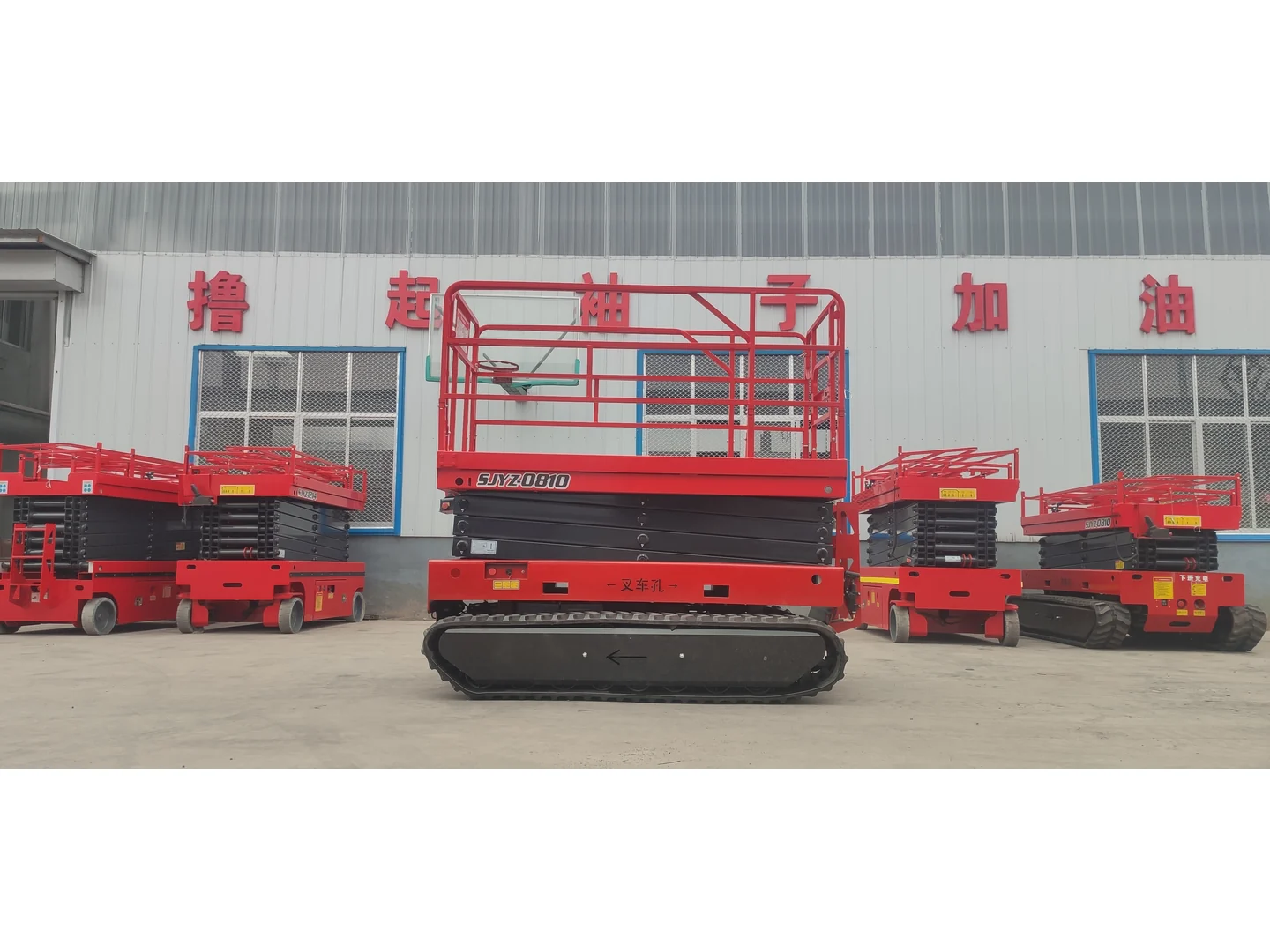 Complete red crawler scissor lift in a lowered front view, with other equipment in the background