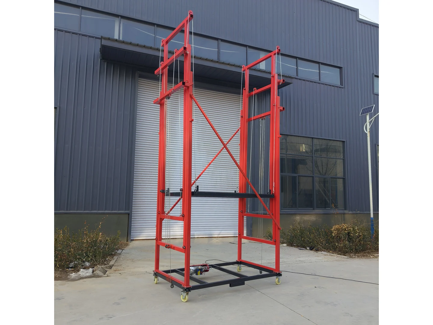 Complete red open-frame electric scaffold beside a building, with platform, pulleys and caster wheels visible